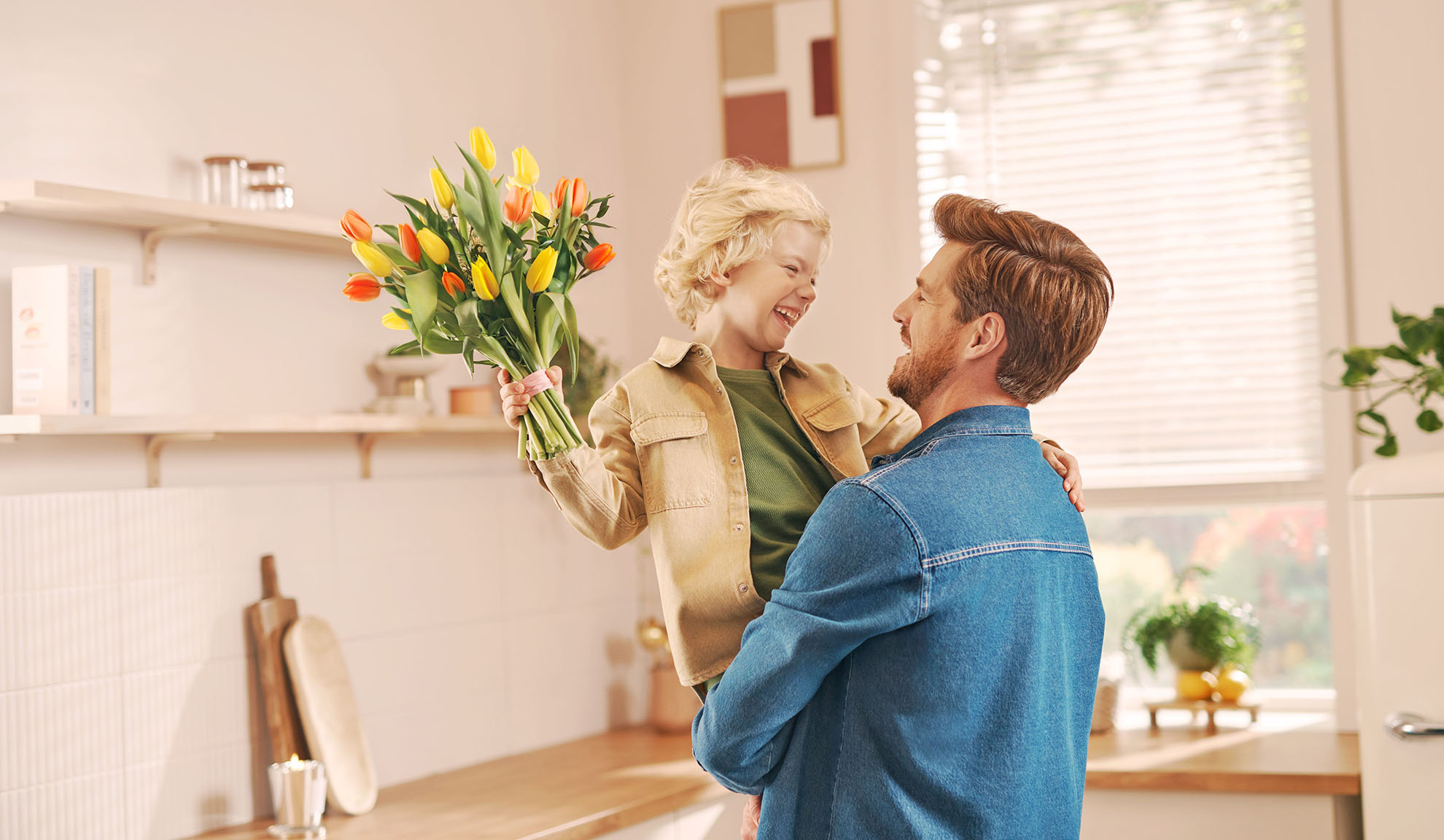 Padre che tiene in braccio il figlio che presenta un mazzo di tulipani in una cucina luminosa.