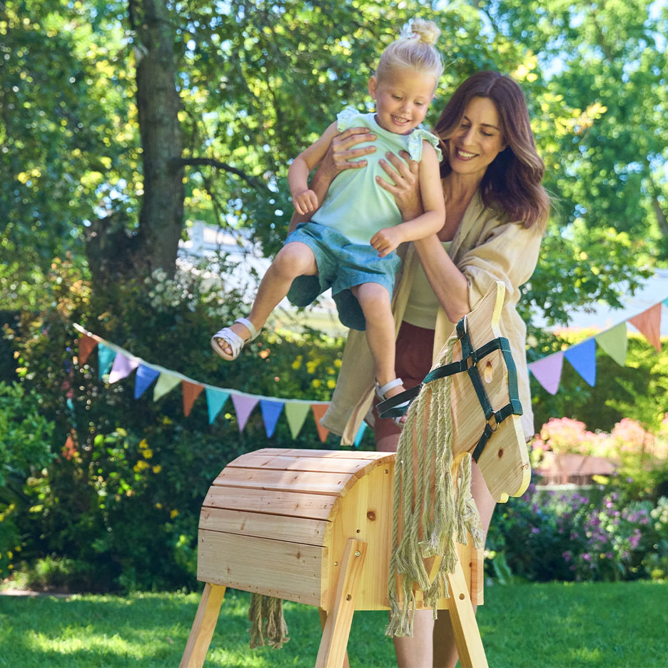 Donna e bambina giocano con un cavallo a dondolo di legno in un giardino con festoni.