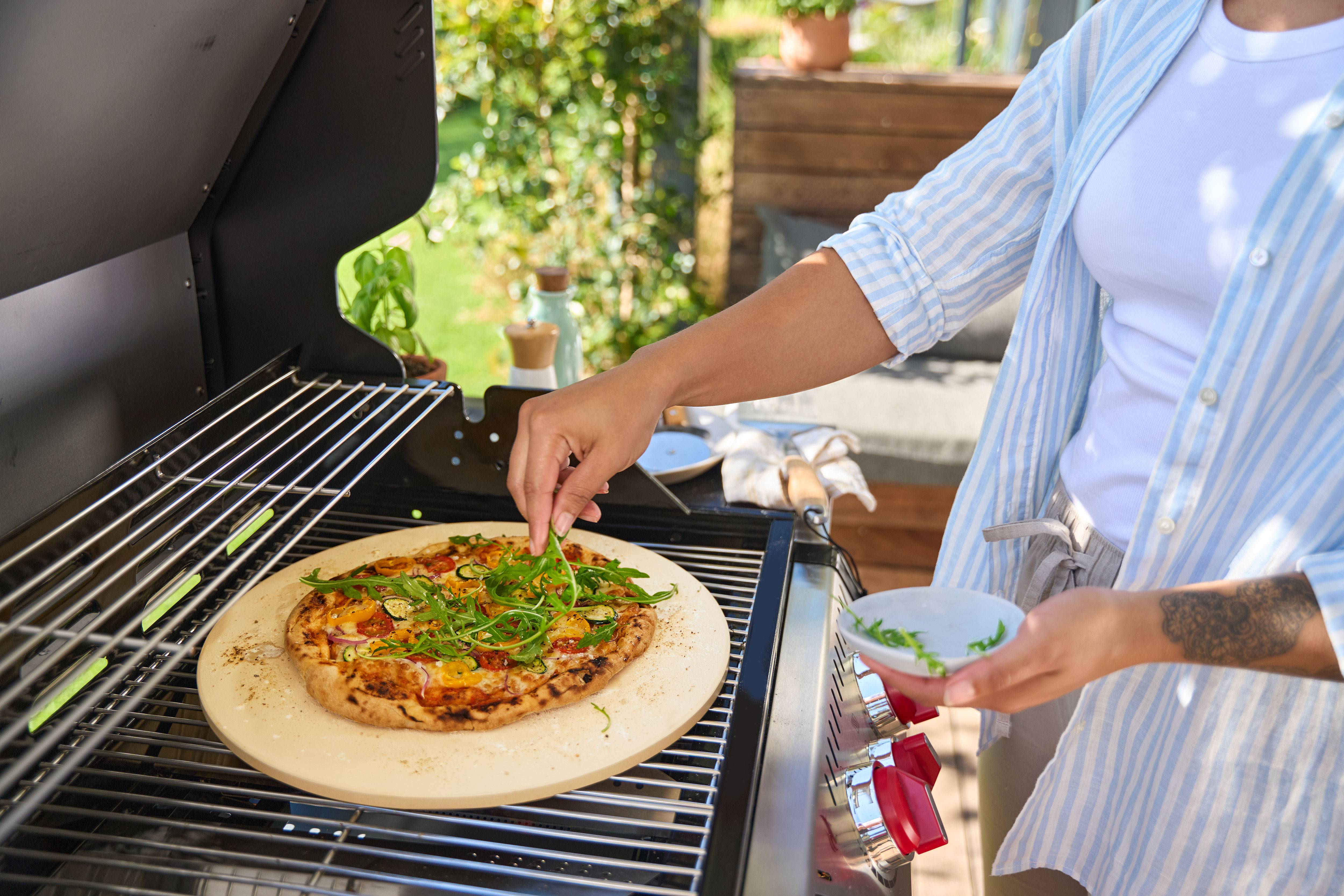 Persona che aggiunge rucola a una pizza su pietra refrattaria in un barbecue a gas.
