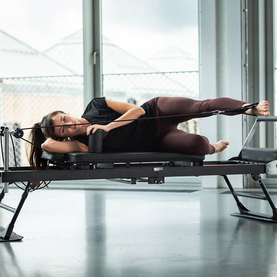 Donna che fa Pilates su una macchina reformer, sdraiata su un fianco con cinghie alle gambe.