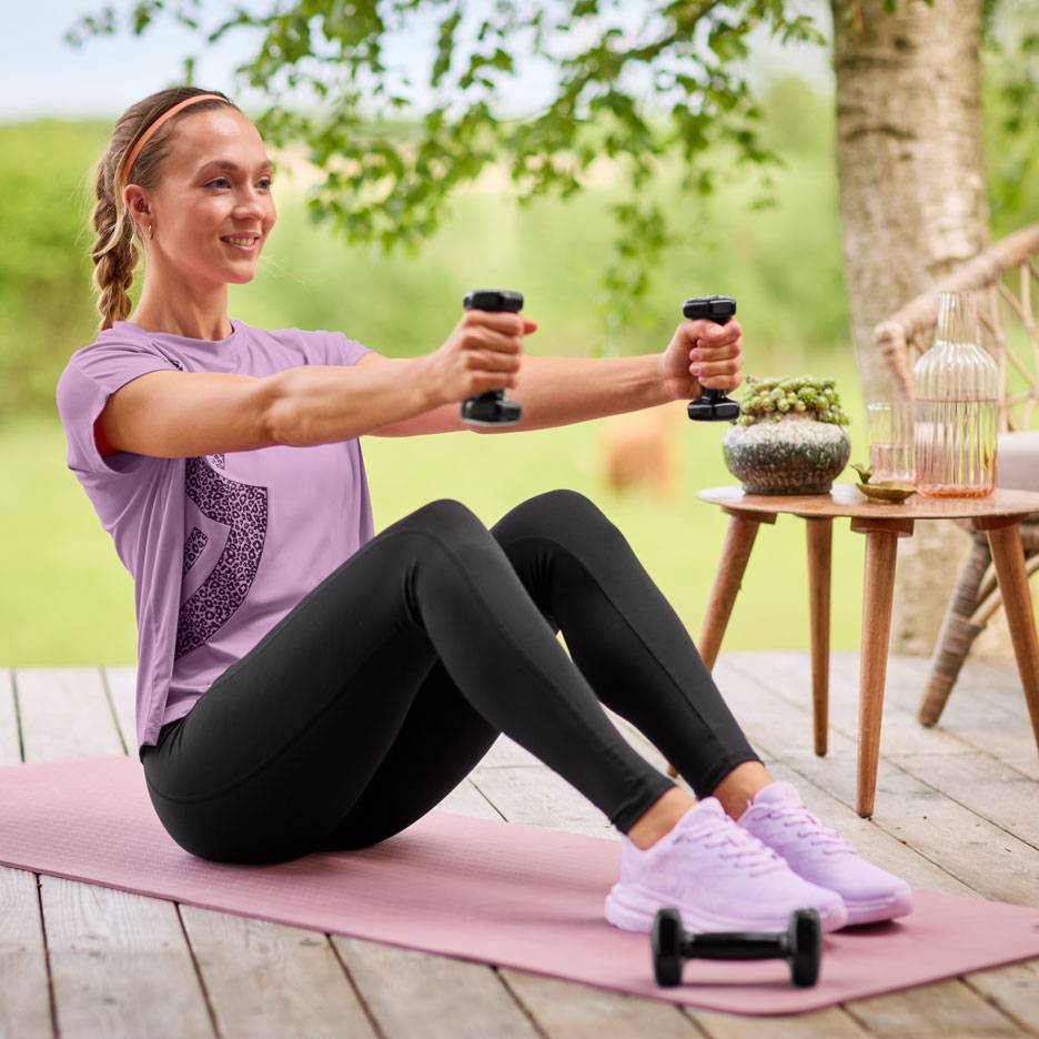 Donna in maglietta viola e leggings neri si allena con manubri su un tappetino da yoga.