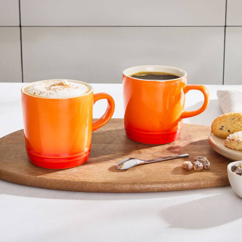 Due tazze da caffè arancioni su un vassoio di legno, una con caffè schiumoso, l'altra con caffè nero.