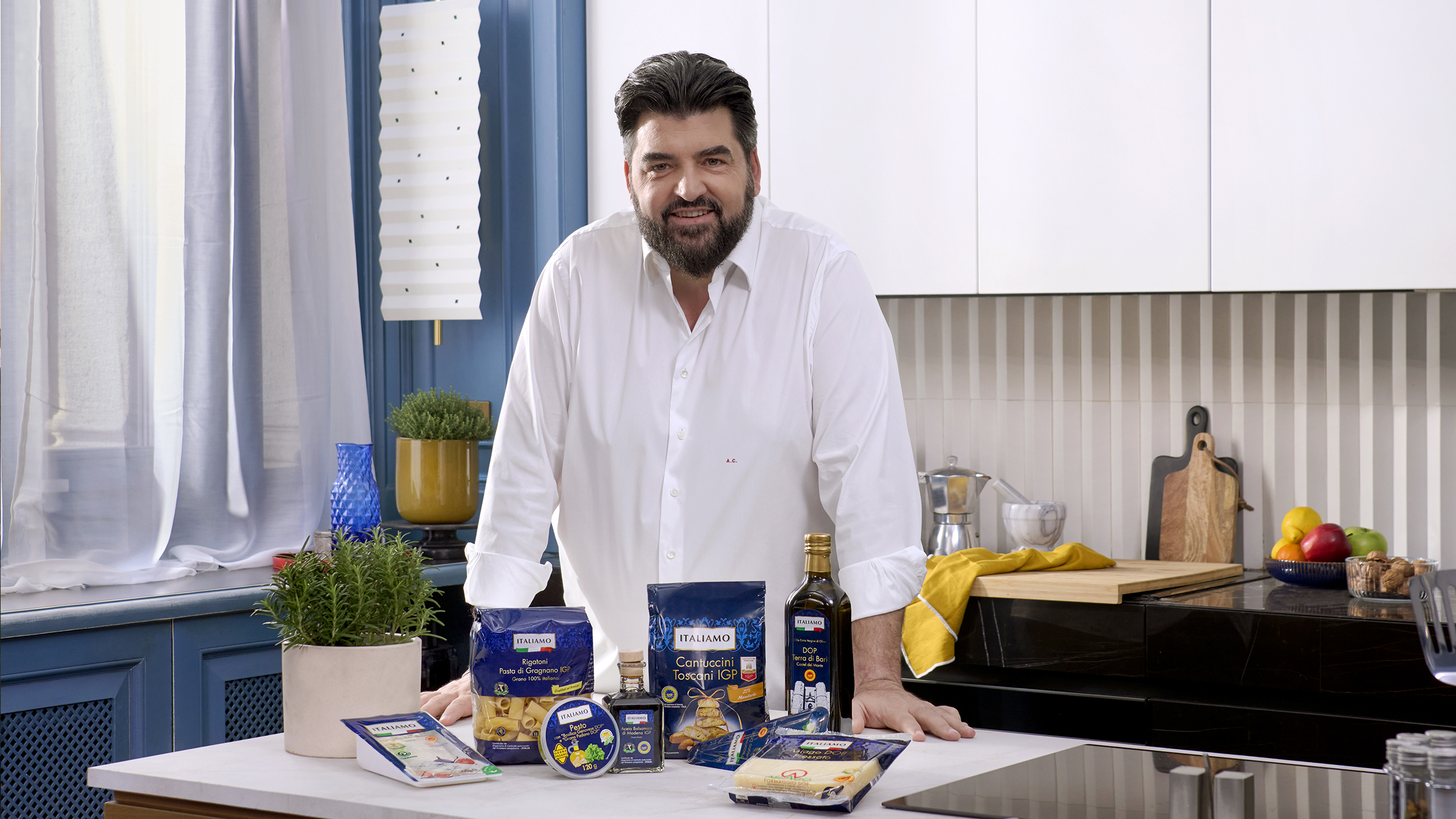 Uomo sorridente in cucina con pasta, pesto, cantuccini, aceto balsamico e olio d'oliva.