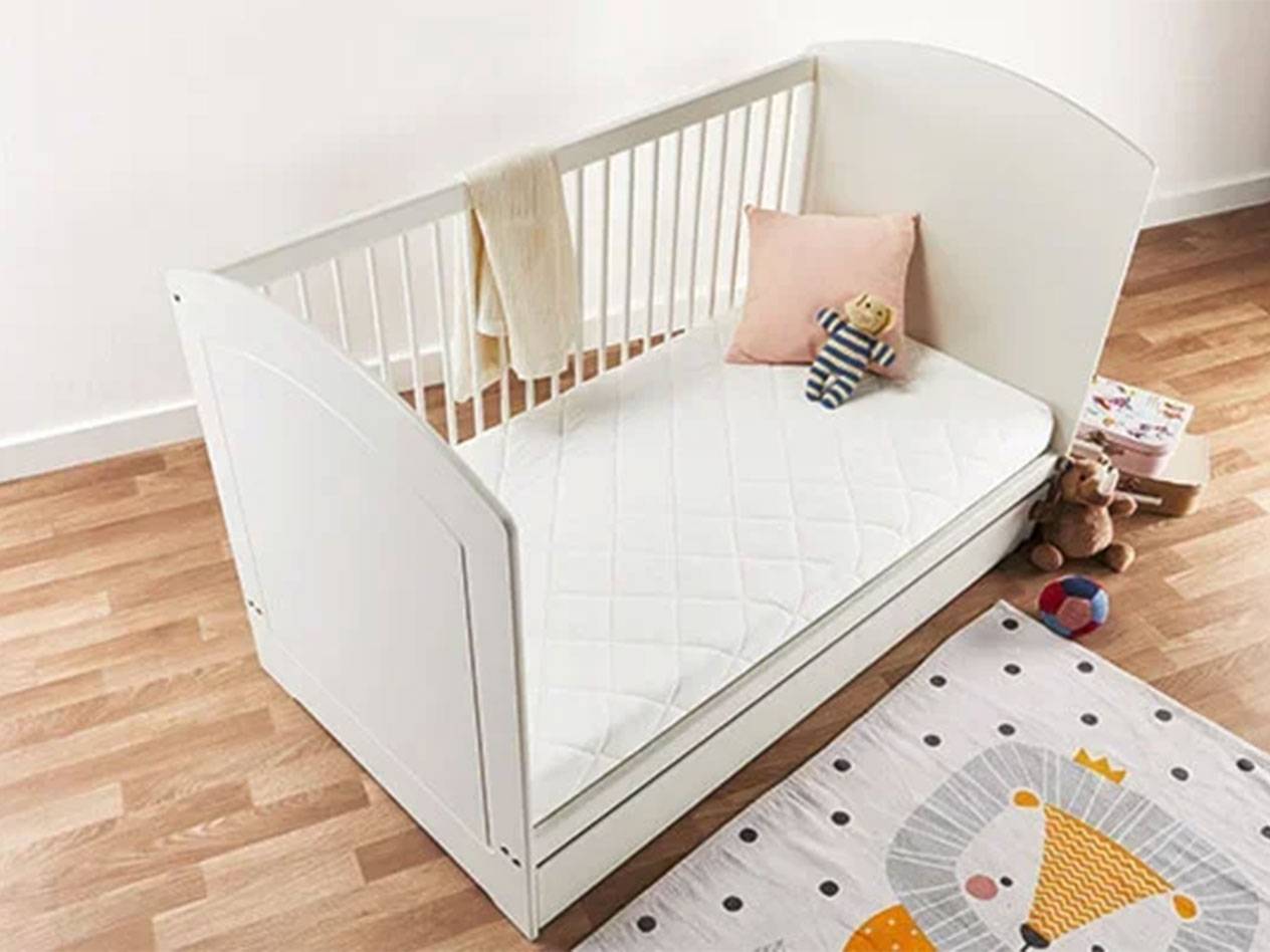 Culla bianca per bambini con materasso, cuscino e giocattoli su pavimento in legno.