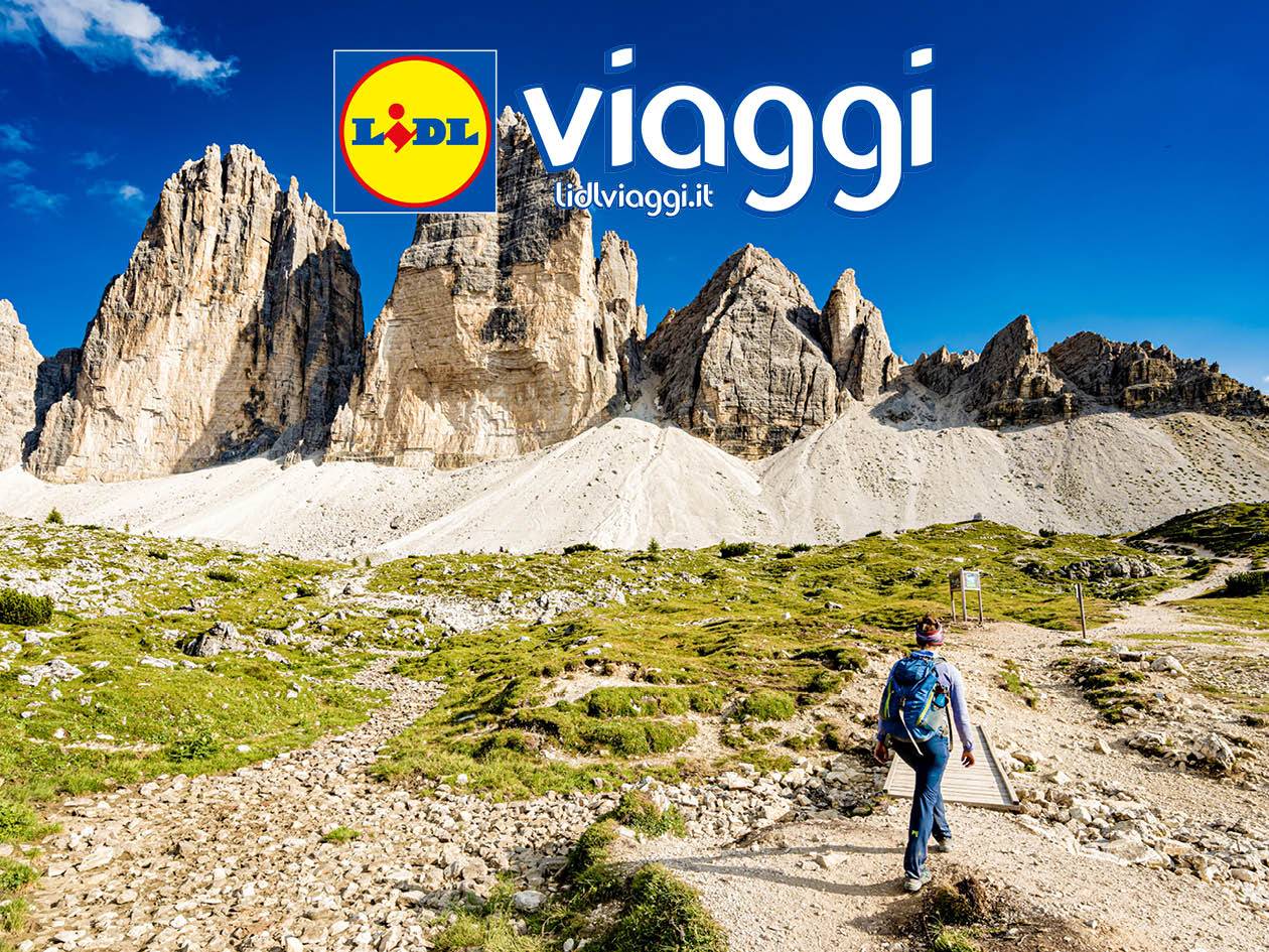 Escursionista con zaino in spalla su un sentiero di montagna, con cime rocciose e cielo blu.