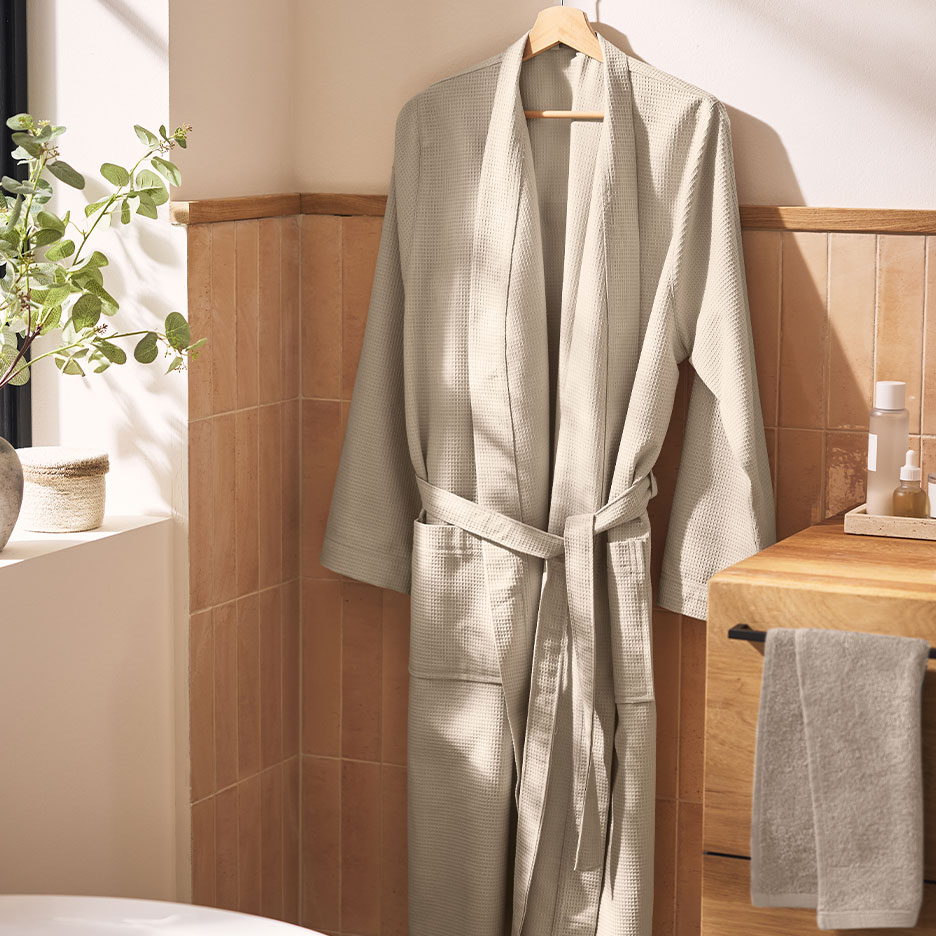 Accappatoio a nido d'ape beige appeso in un bagno con piastrelle marroni e piante.