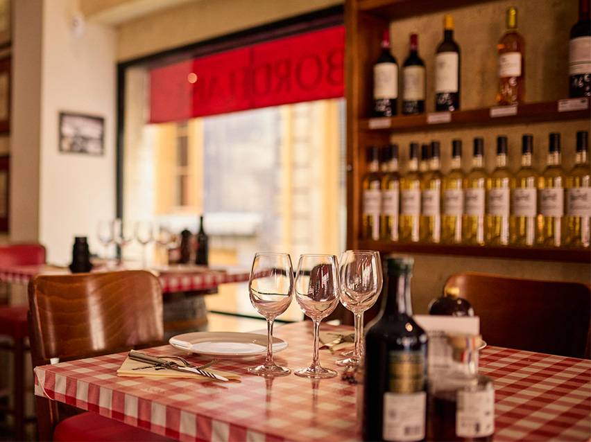 Tavolo da ristorante con tovaglia a quadri rossi e bianchi, calici da vino e bottiglie di vino.