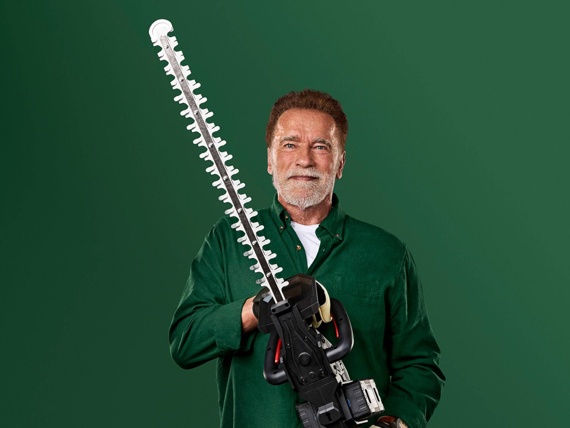 Arnold Schwarzenegger tiene un grande tagliasiepi su uno sfondo verde.