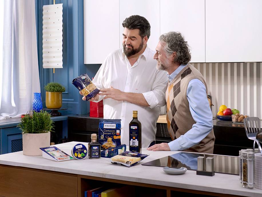 Due uomini in cucina con pasta, biscotti, aceto balsamico e formaggio sul bancone.