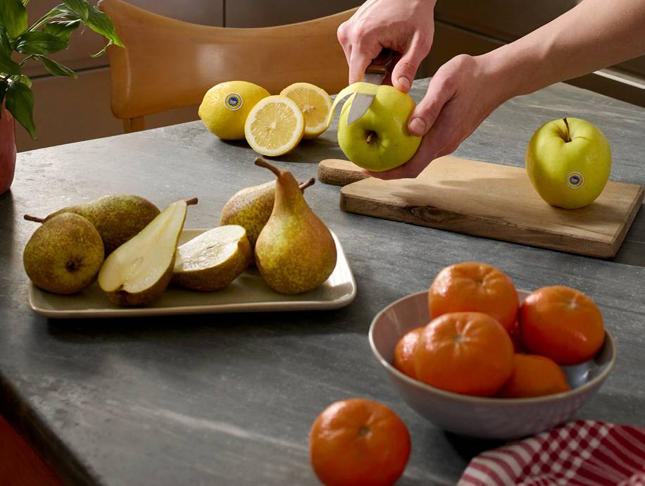 Mani che sbucciano una mela verde su un tagliere, con pere, limoni e mandarini sul tavolo.