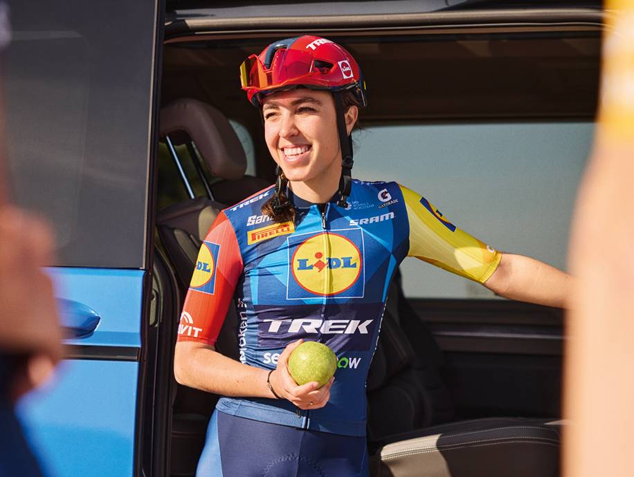 Ciclista donna sorridente con casco e maglia sportiva, tiene una mela verde, accanto a un veicolo.