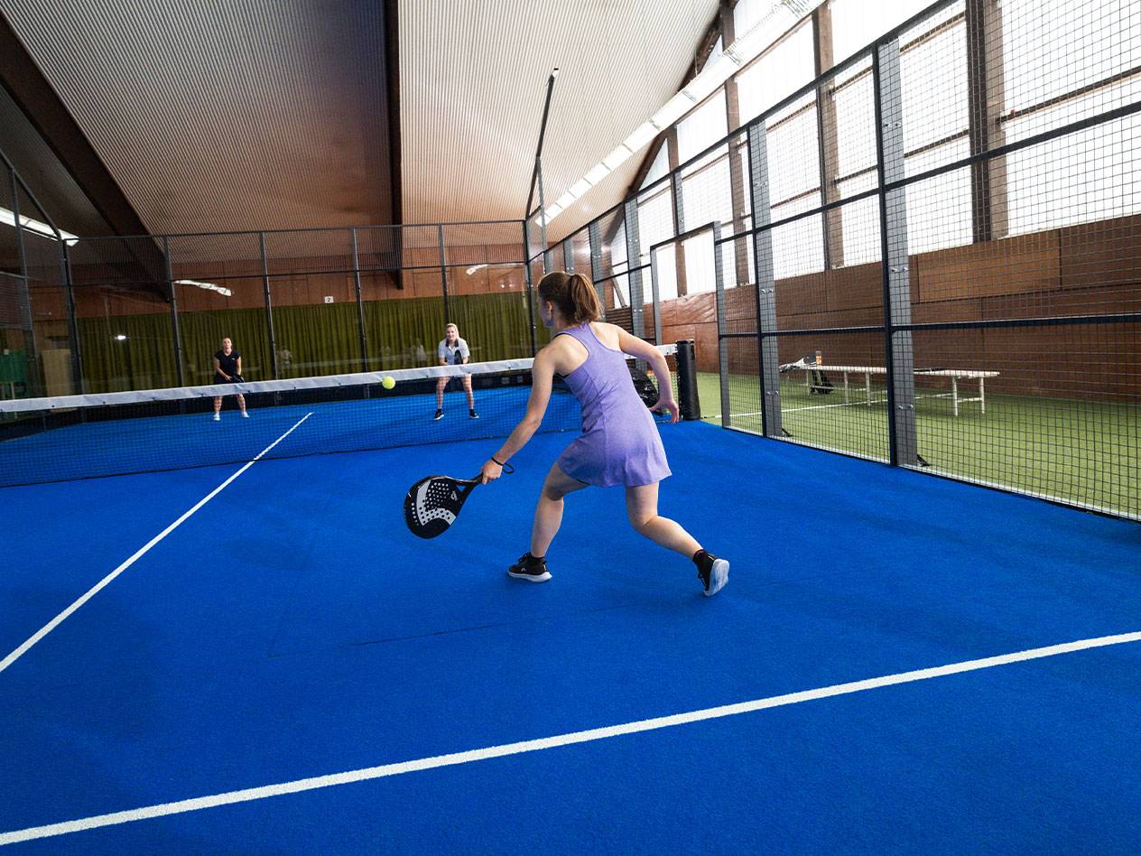 Donna che gioca a padel su un campo blu, con altri giocatori sullo sfondo.