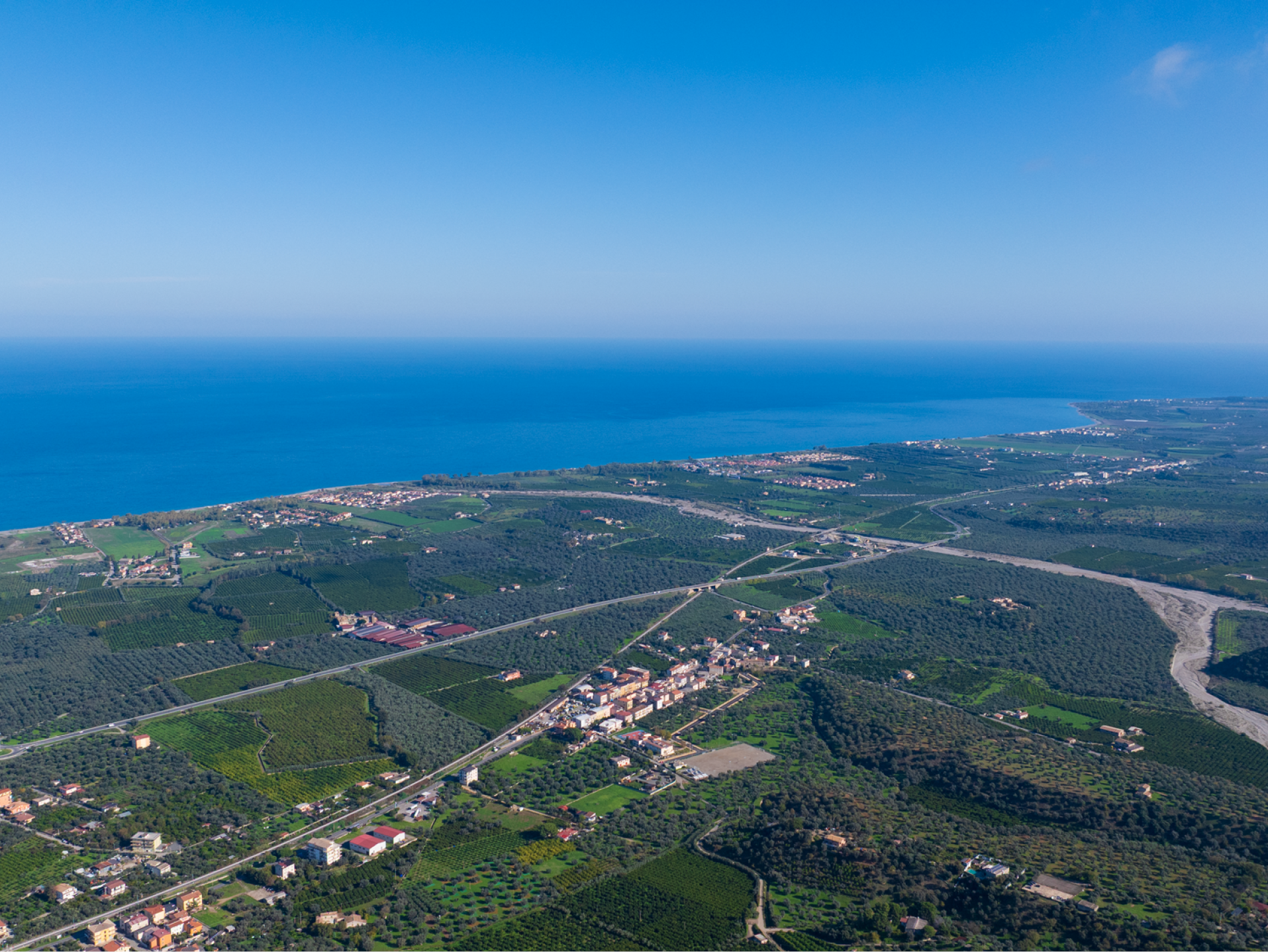Vista aerea di un paesaggio costiero con campi verdi, alberi e un piccolo centro abitato.
