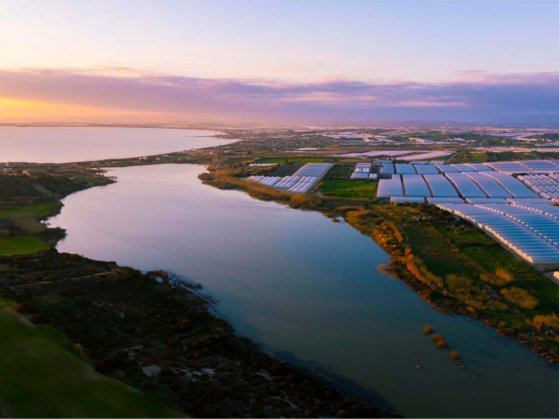 Serre agricole e un lago al tramonto, con il cielo che sfuma dal viola all'arancione.