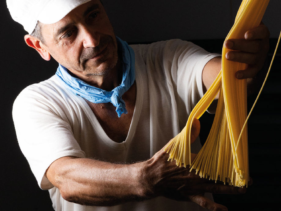 Uomo con cappello bianco e bandana blu che tiene in mano spaghetti crudi.
