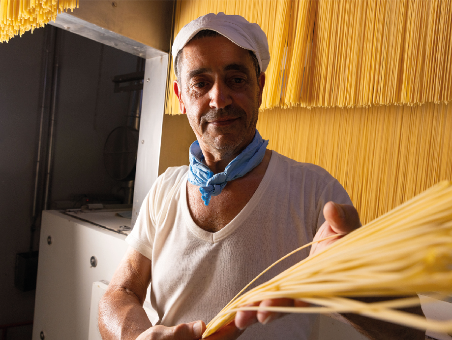 Uomo con cappello e bandana blu che tiene in mano spaghetti freschi, con spaghetti appesi sullo sfondo.