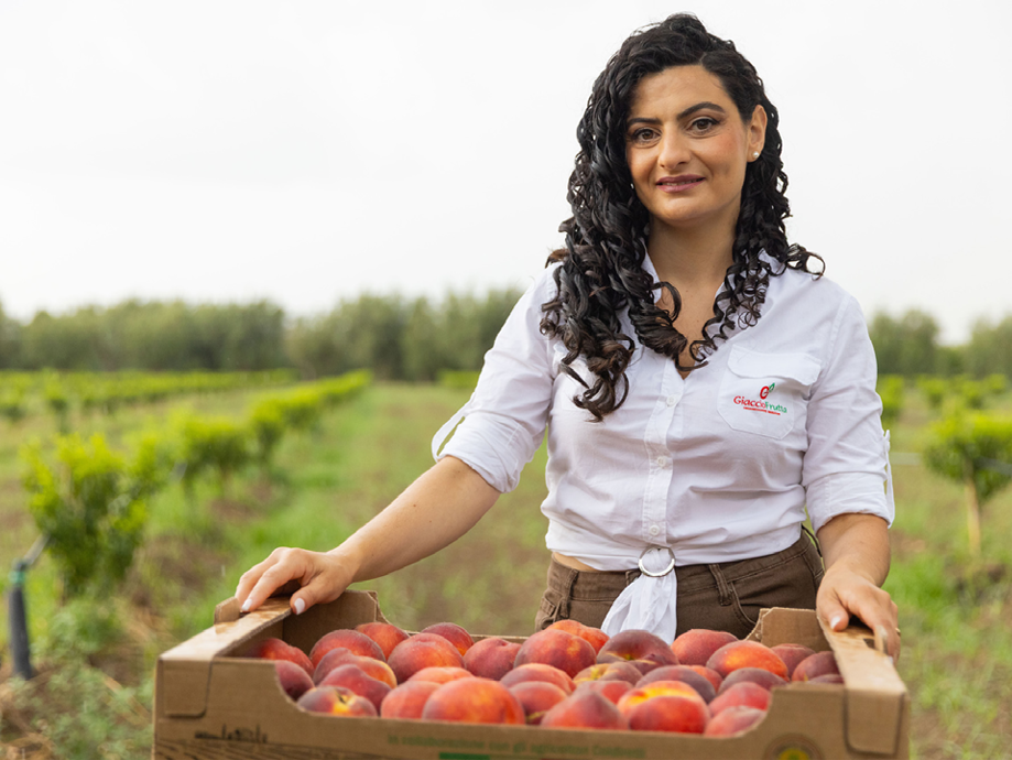 Donna sorridente in un campo con una cassetta di pesche fresche.