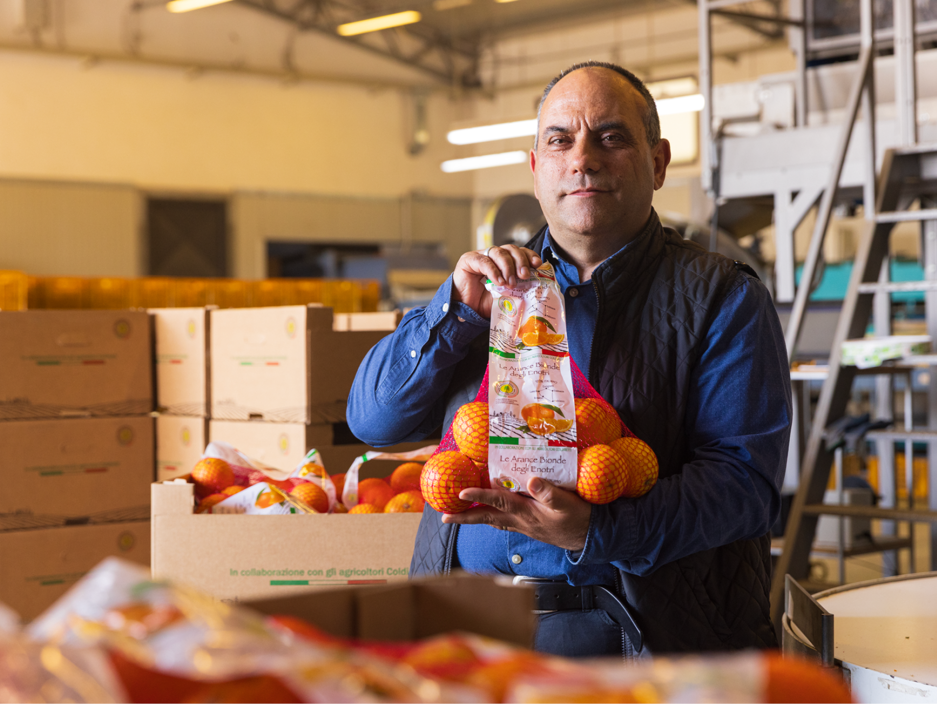 Un uomo in un magazzino tiene in mano una confezione di arance, con altre scatole sullo sfondo.