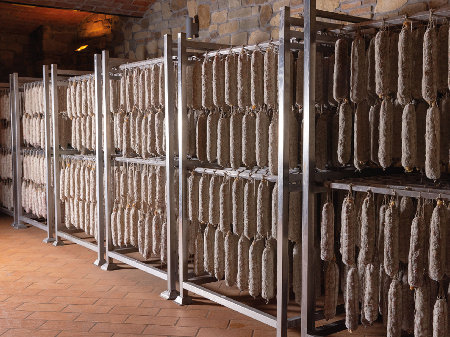 File di salami stagionati appesi su rastrelliere metalliche in una cantina in pietra.