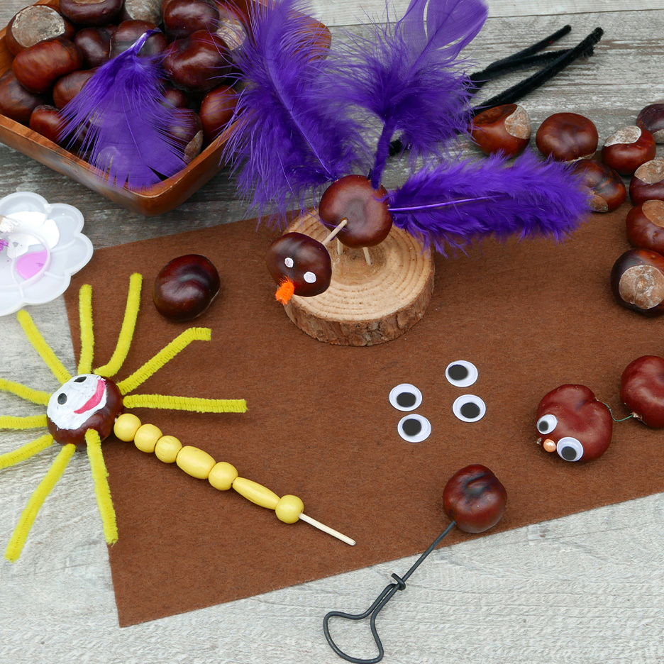 Artigianato di castagne con piume viola, occhi mobili e fili di ciniglia su un tavolo di legno.
