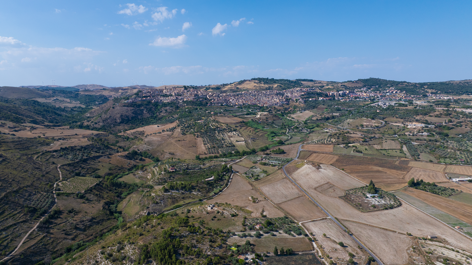 Vista aerea di un paesaggio rurale con campi, colline e un paese in lontananza.