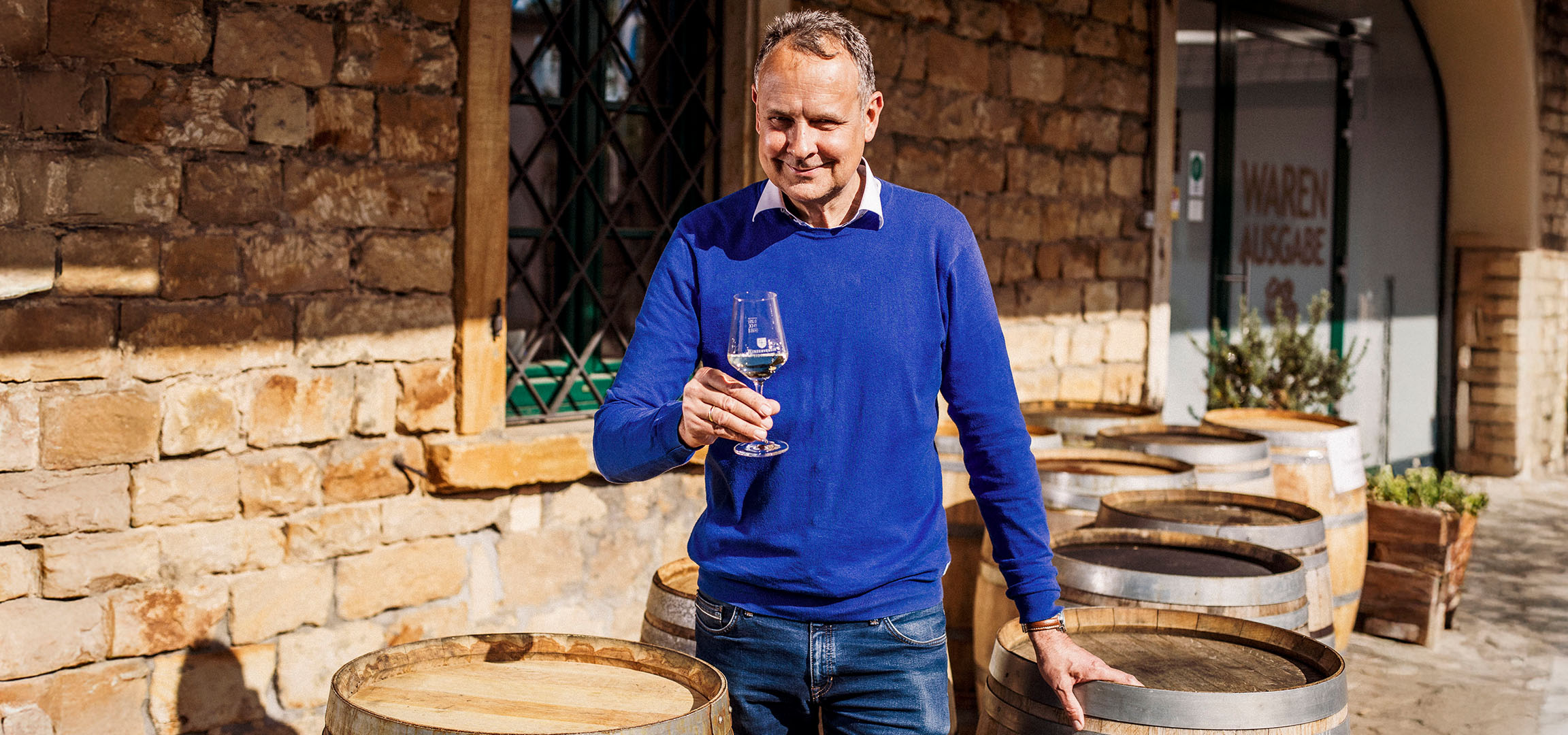 Uomo sorridente in maglione blu che tiene un bicchiere di vino bianco davanti a un muro di pietra.