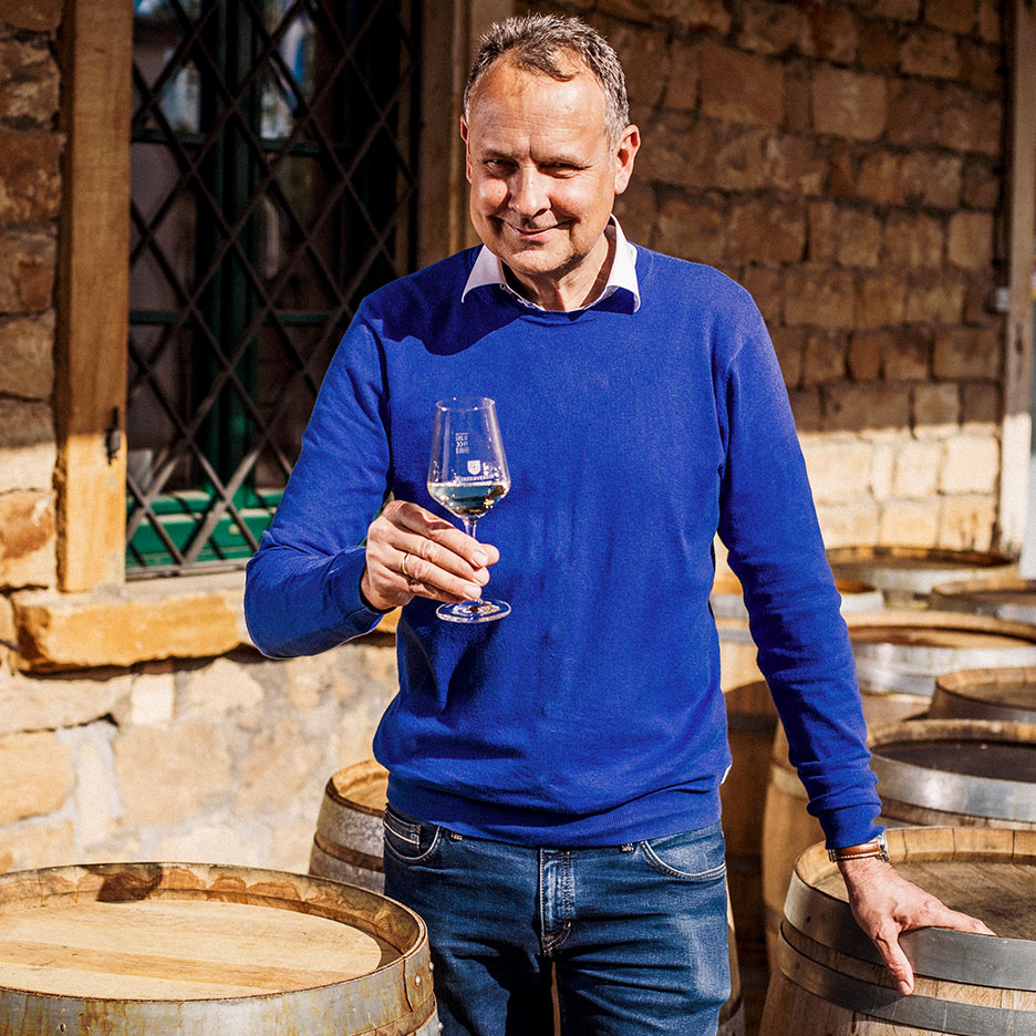 Uomo sorridente in maglione blu che tiene un bicchiere di vino, con botti di legno sullo sfondo.