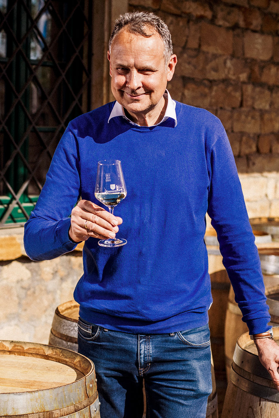 Uomo sorridente in maglione blu e jeans, tiene un bicchiere di vino bianco tra botti di legno.
