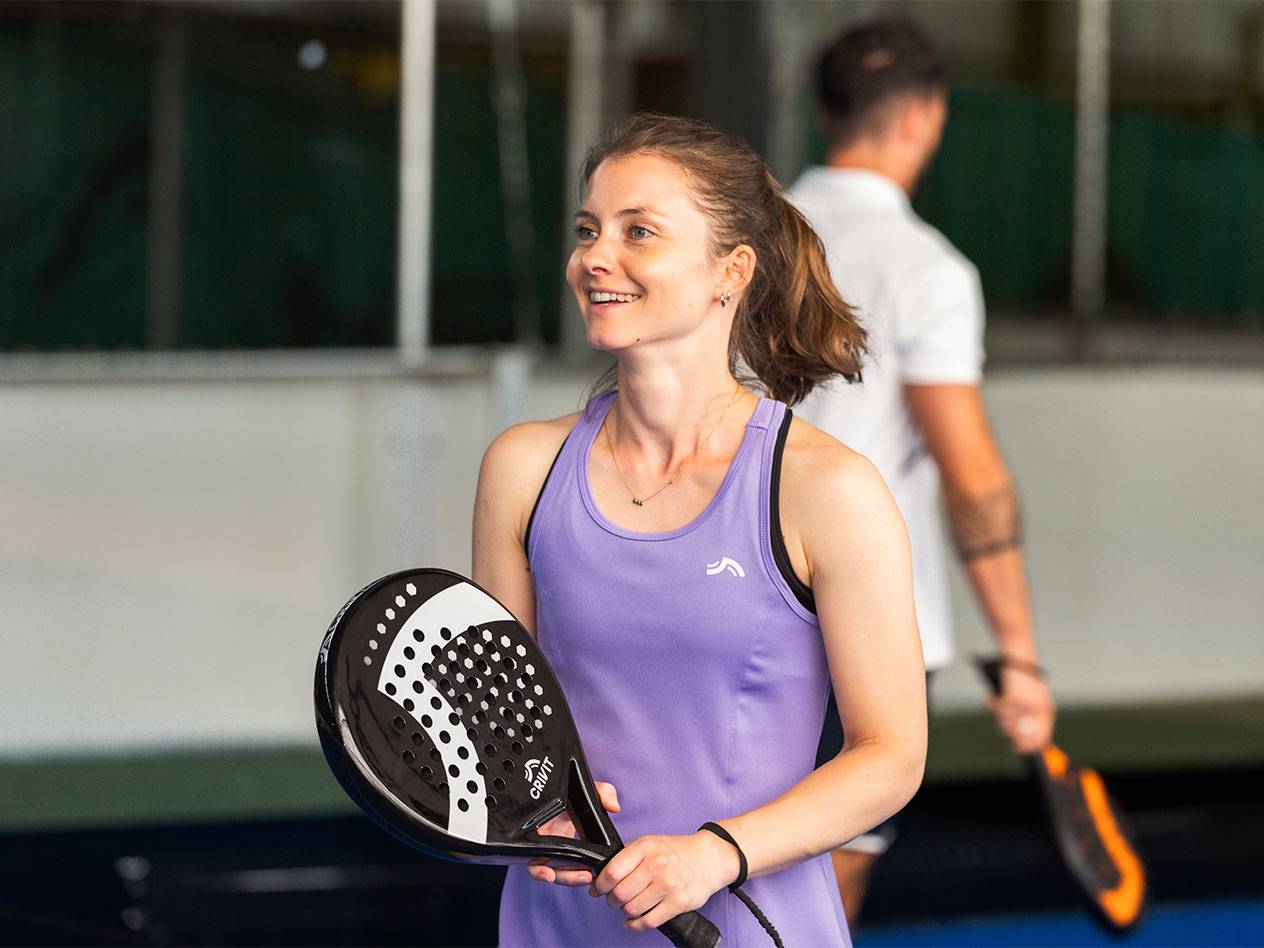 Donna in abito sportivo viola che tiene una racchetta da padel, uomo sullo sfondo.