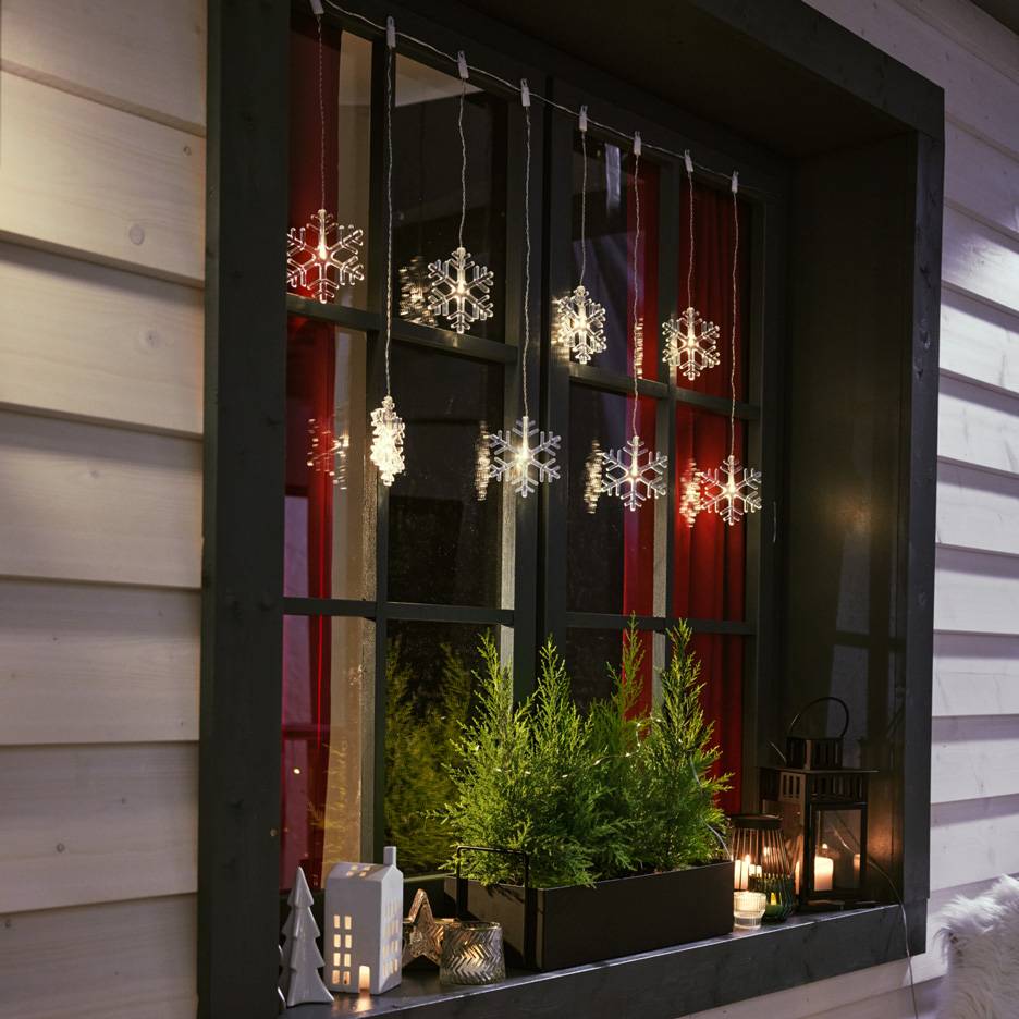 Luci a fiocco di neve appese a una finestra con piccoli abeti e candele sul davanzale.