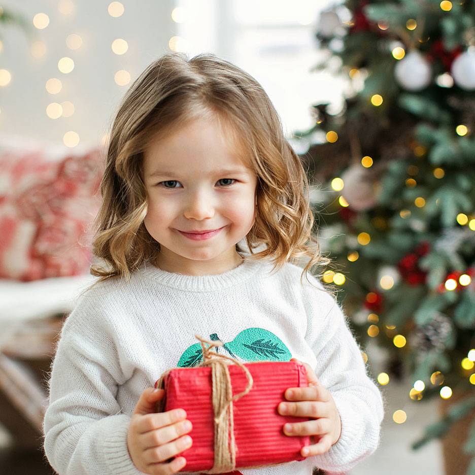Bambina sorridente con un regalo di Natale, con un albero di Natale illuminato sullo sfondo.