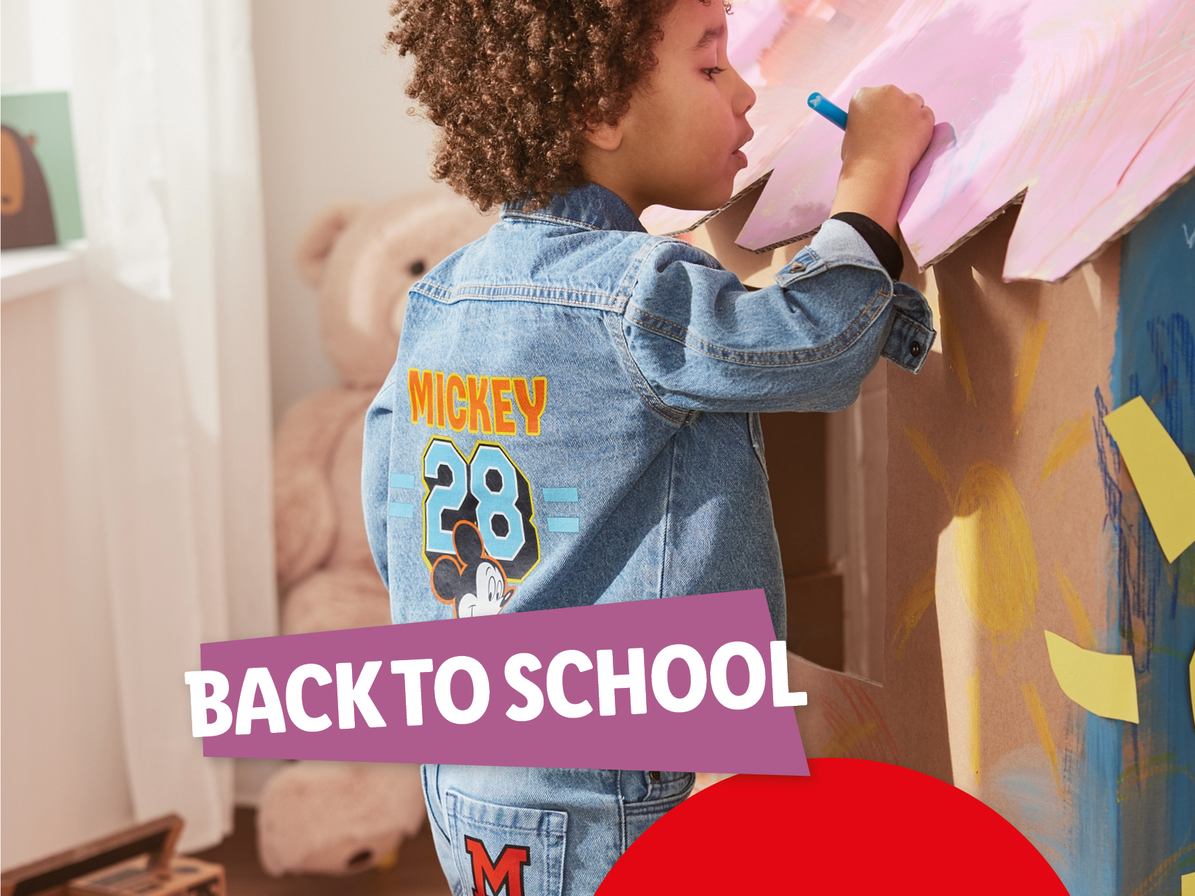 Un bambino con una giacca di jeans di Topolino disegna su una scatola di cartone, con la scritta 'BACK TO SCHOOL'.