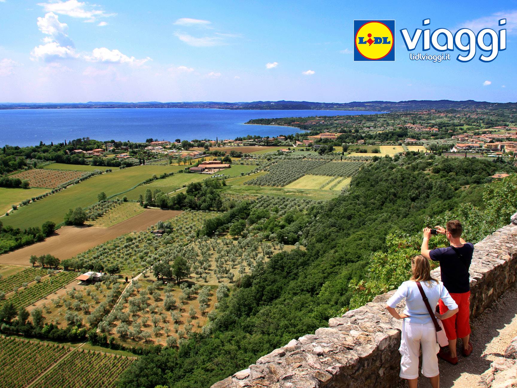 Due persone ammirano un paesaggio con un lago, campi e alberi, con il logo Lidl Viaggi.