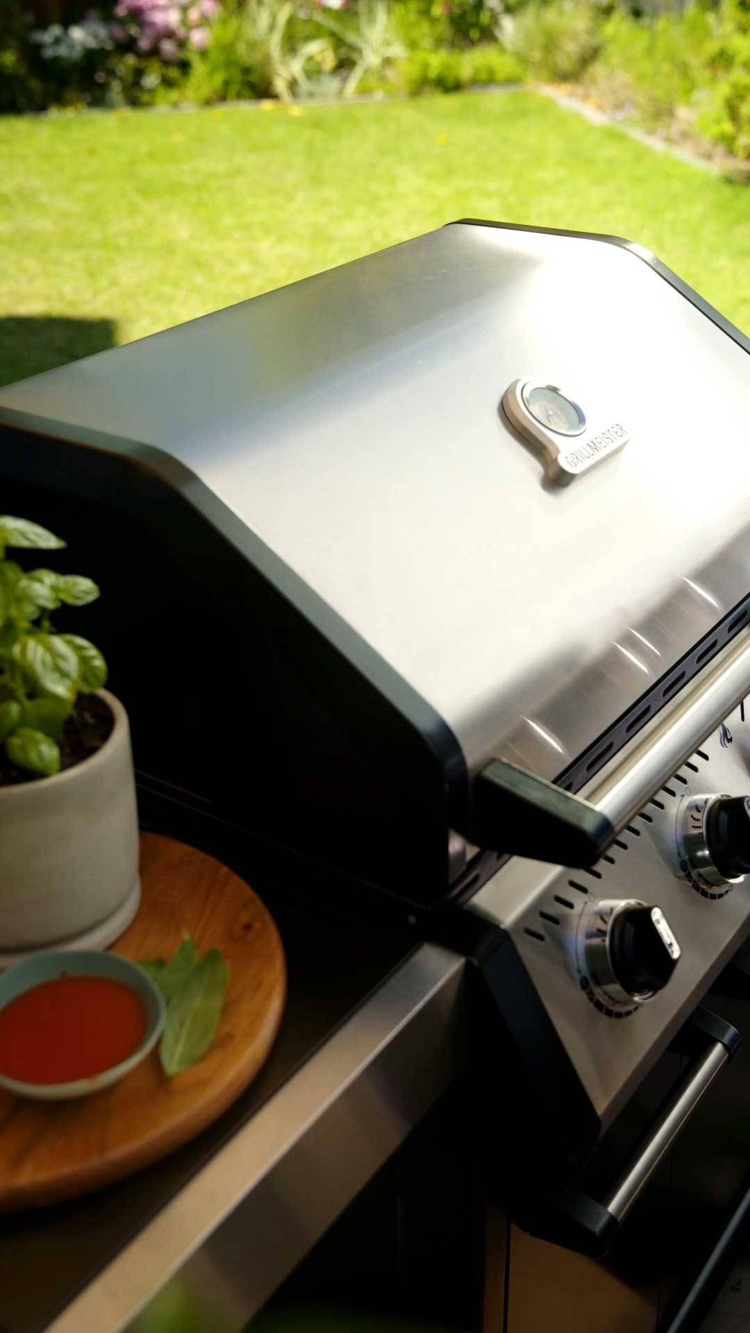 Barbecue a gas in acciaio inossidabile con termometro, accanto a un vaso di basilico e salsa.