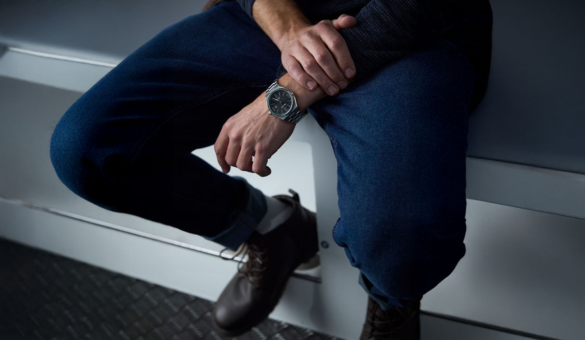 Uomo con jeans blu scuro, maglione scuro e orologio d'argento, seduto.