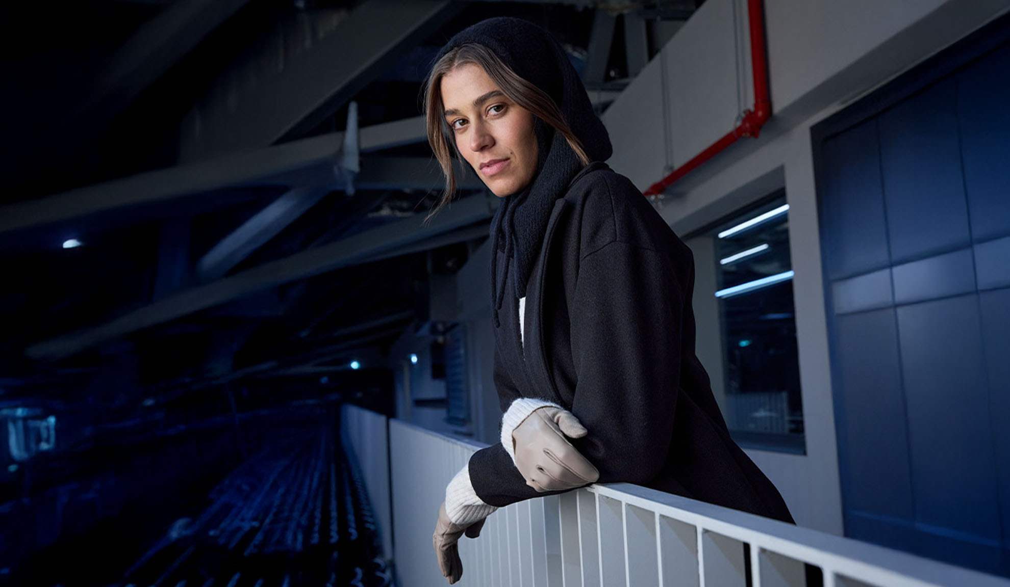 Donna in cappotto nero e sciarpa con cappuccio, appoggiata a una ringhiera in un ambiente moderno.
