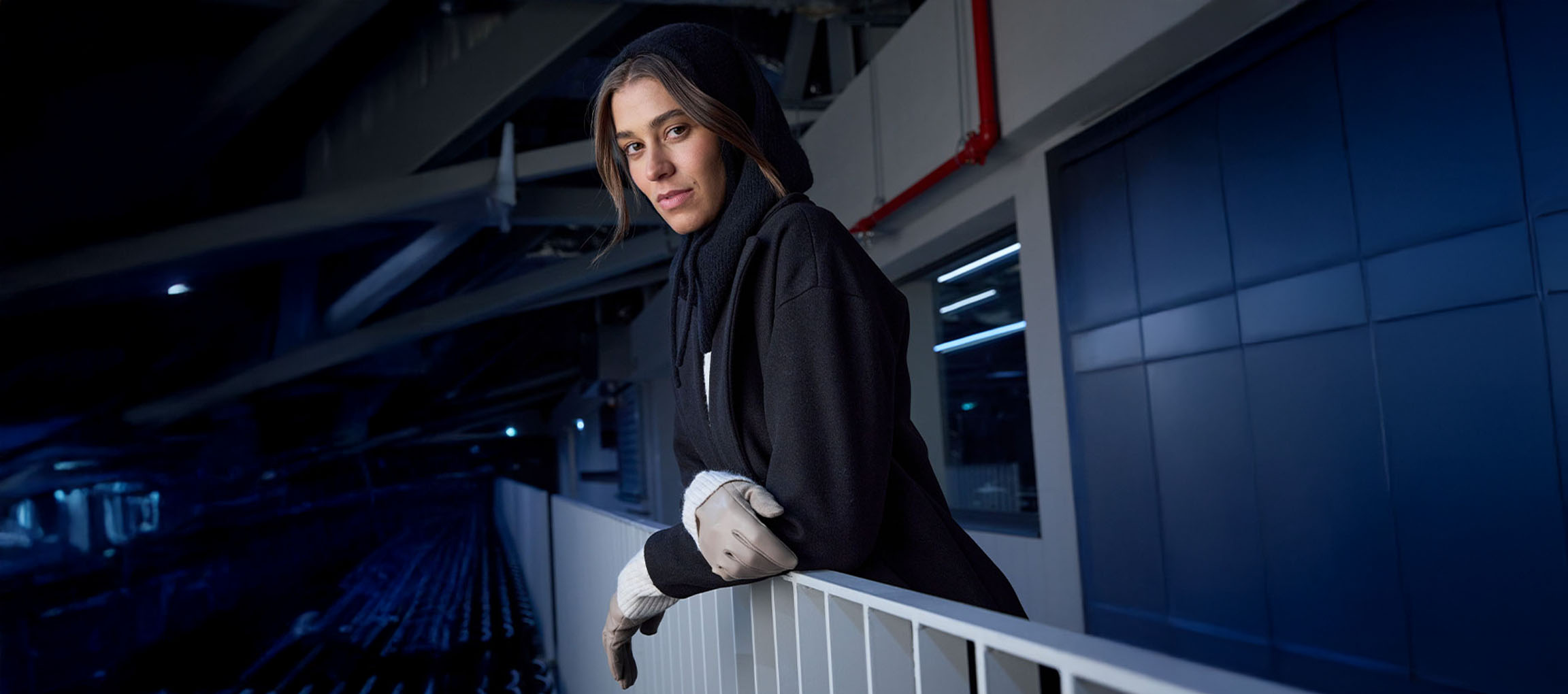 Donna in cappotto nero e guanti appoggiata a una ringhiera in ambiente urbano.