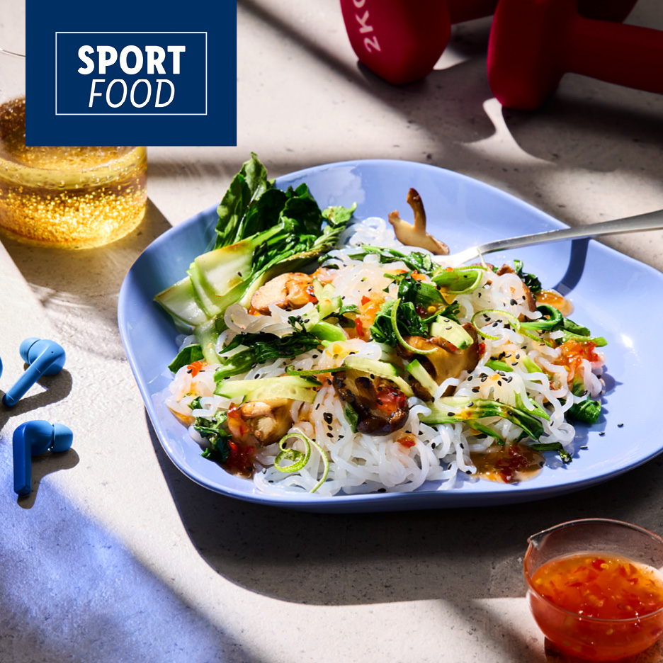 Piatto di noodles con verdure e gamberi, cuffie e manubri sullo sfondo.