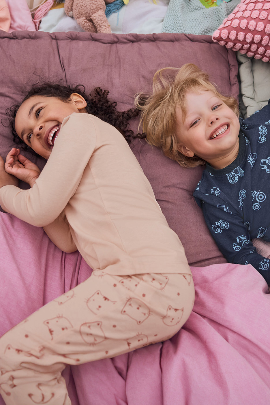 Due bambini sorridenti in pigiama, uno con gatti e l'altro con trattori, sdraiati su un letto.