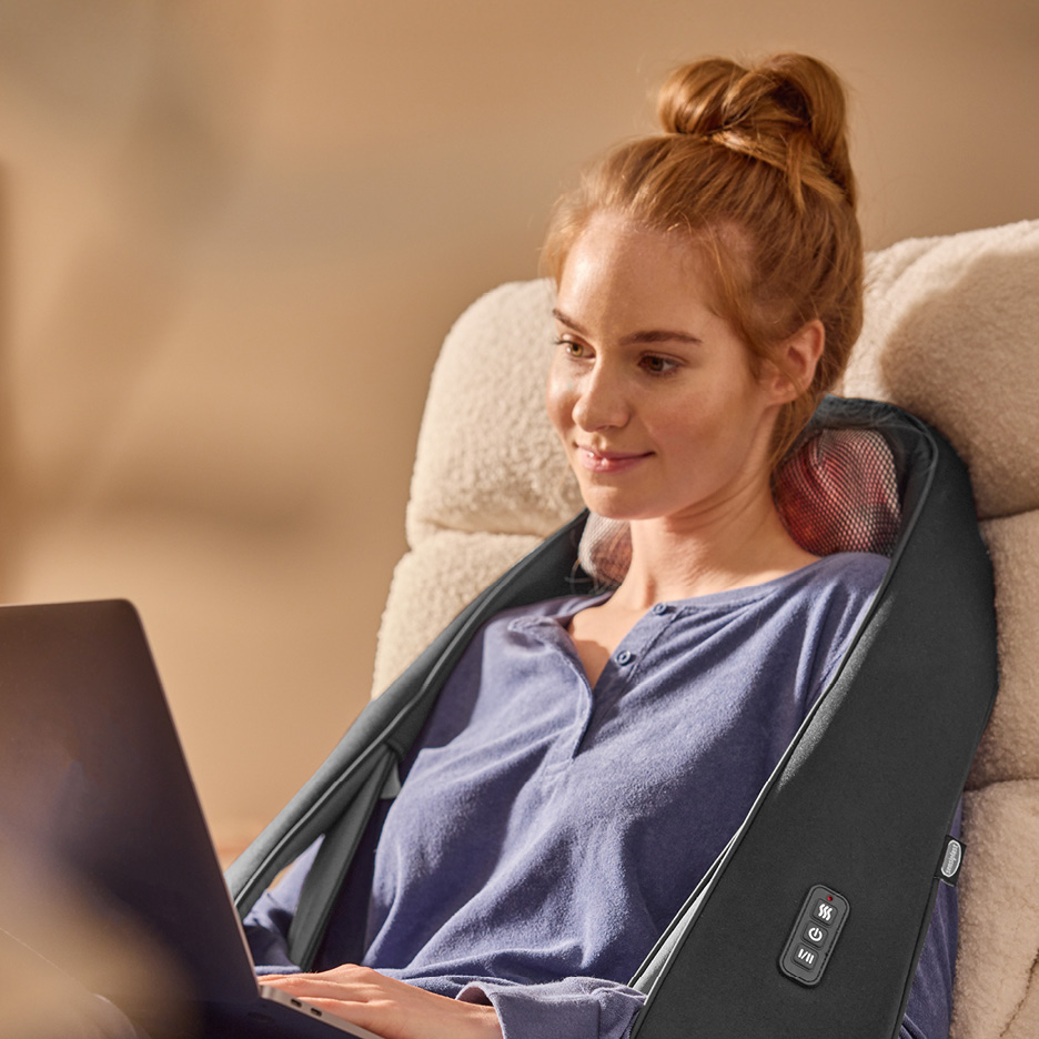 Donna con massaggiatore cervicale e dorsale, seduta su una poltrona, usa il laptop.