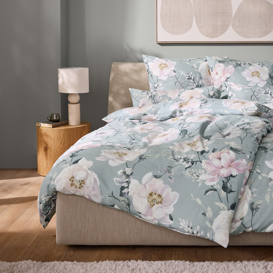 Letto con copripiumino floreale verde acqua, comodino in legno e lampada da tavolo.