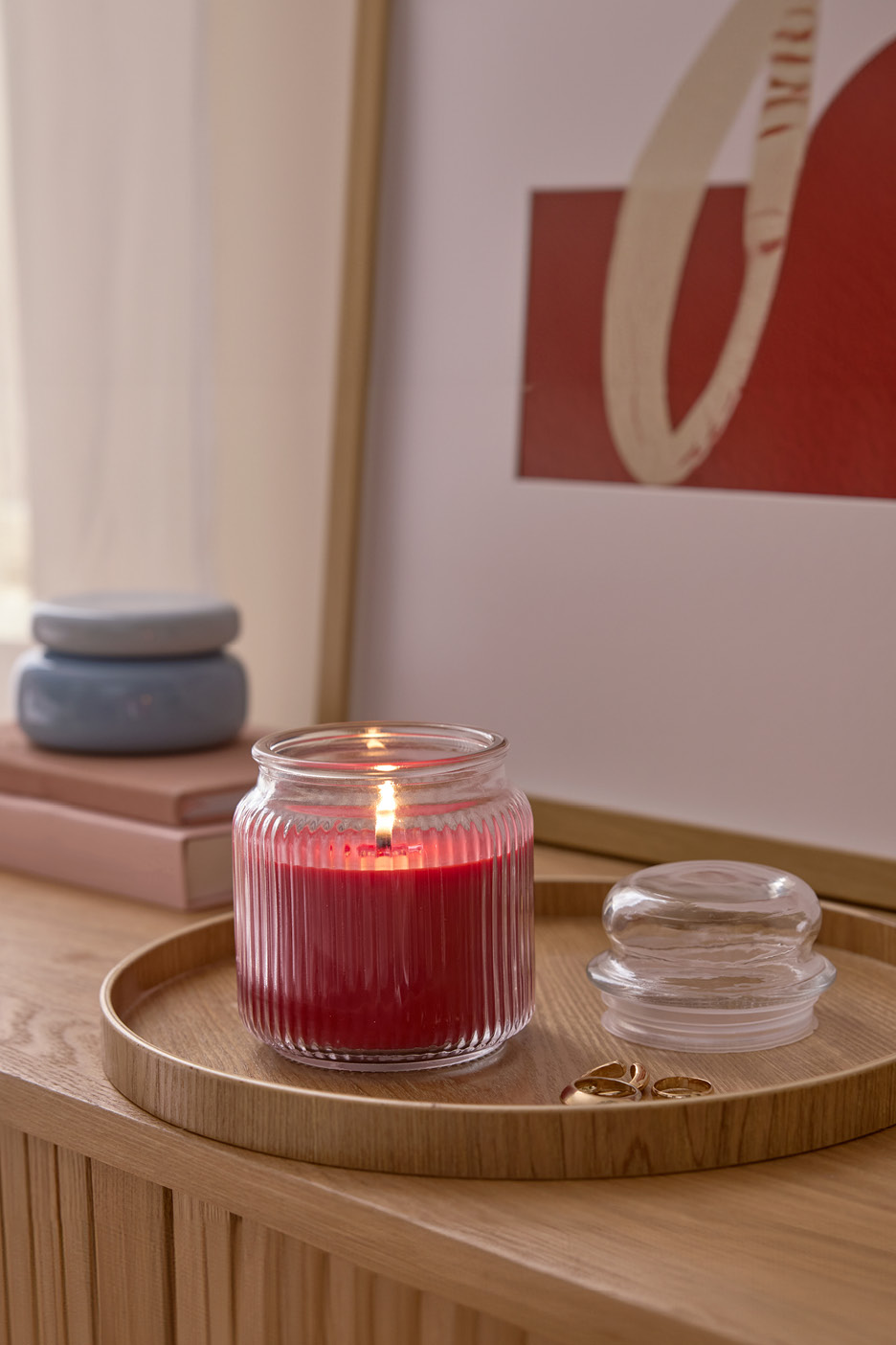 Candela rossa accesa su vassoio di legno con coperchio di vetro e anelli dorati, sullo sfondo libri e un quadro.