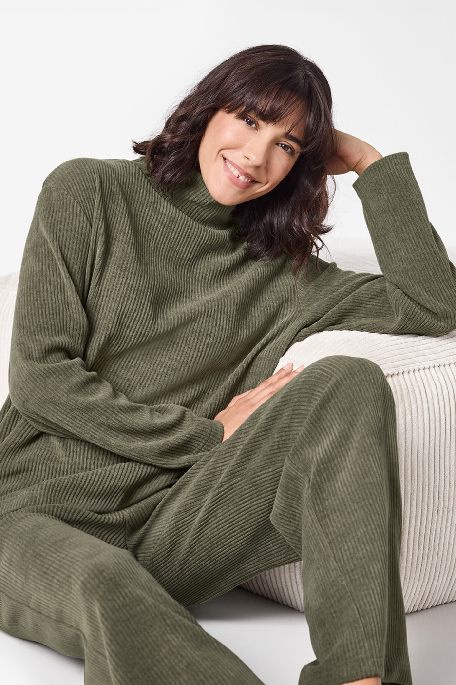 Donna sorridente in completo loungewear a coste verde oliva, seduta su un pouf bianco.