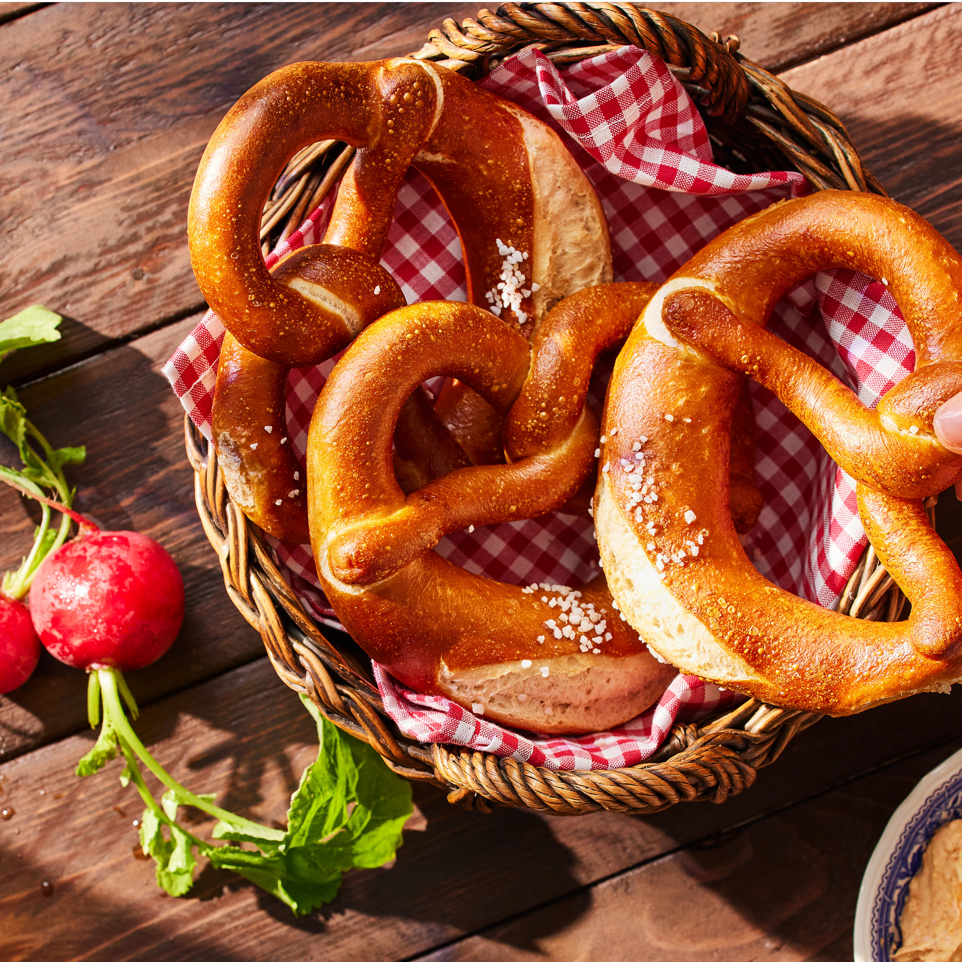 Cesto di vimini con pretzel salati e un panno a quadri rossi, con ravanelli freschi e una ciotola di formaggio spalmabile su un tavolo di legno.