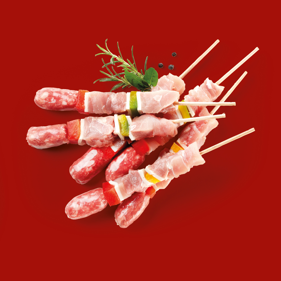 Spiedini di carne cruda e salsicce con verdure e rosmarino su sfondo rosso.