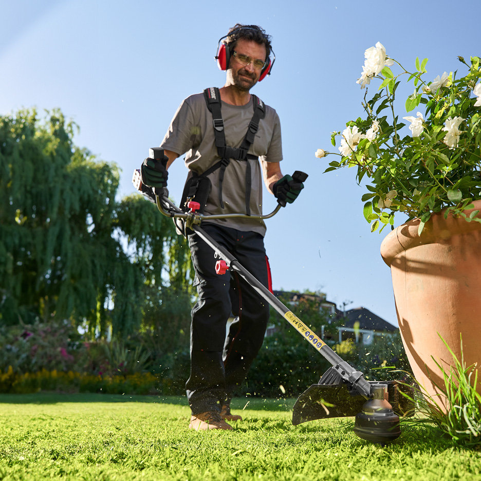 Uomo con decespugliatore a batteria e cuffie antirumore che taglia l'erba in un giardino.