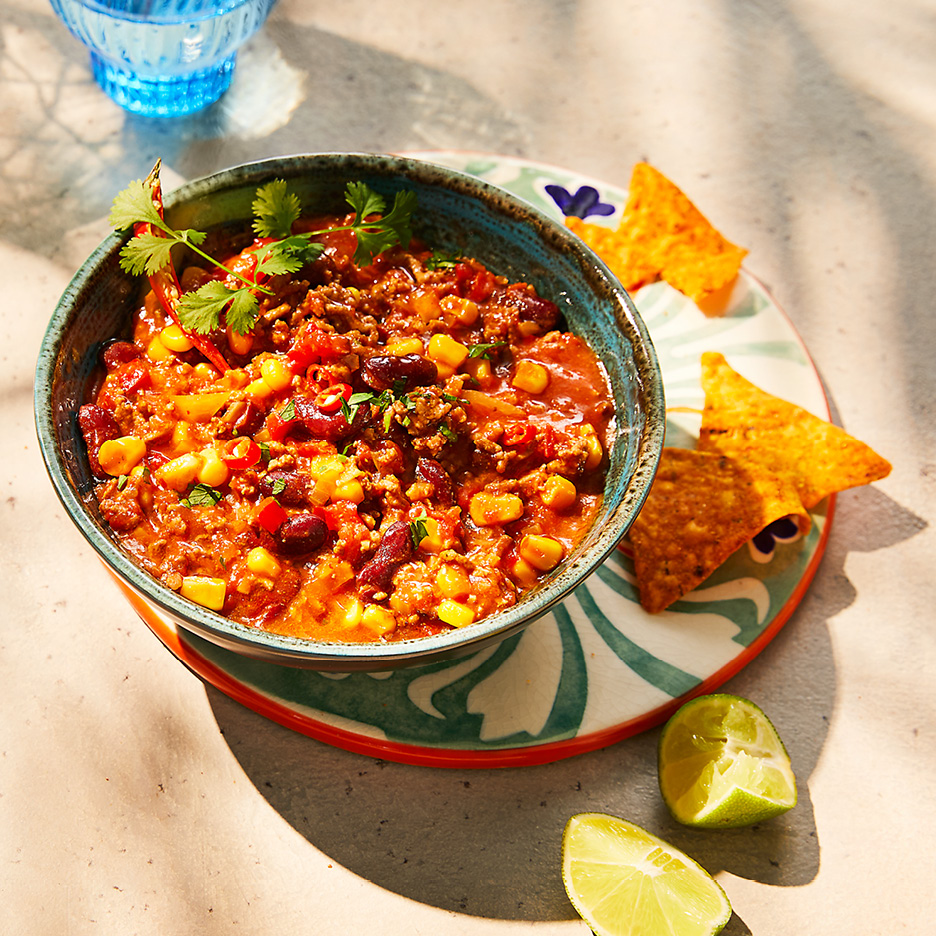 Ciotola di chili con carne con mais, fagioli e coriandolo, accompagnata da nachos e lime.