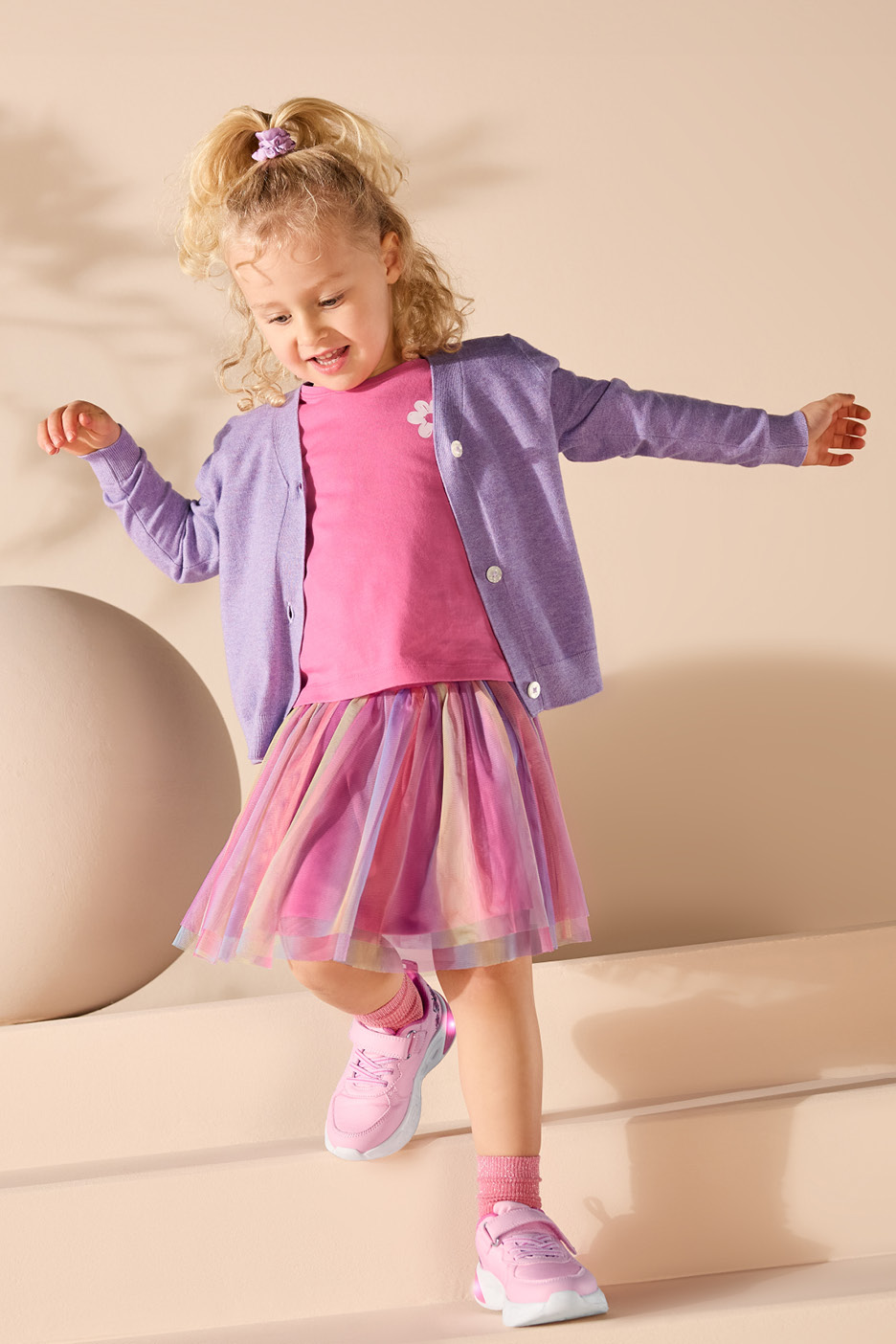 Bambina con cardigan lilla, maglietta rosa, gonna in tulle colorata e scarpe da ginnastica rosa.