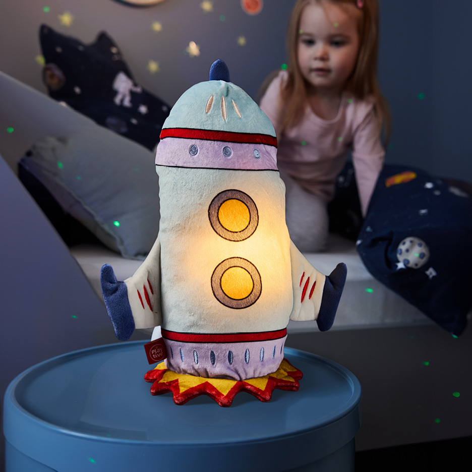 Lampada notturna a forma di razzo su un tavolino blu, con una bambina sullo sfondo.