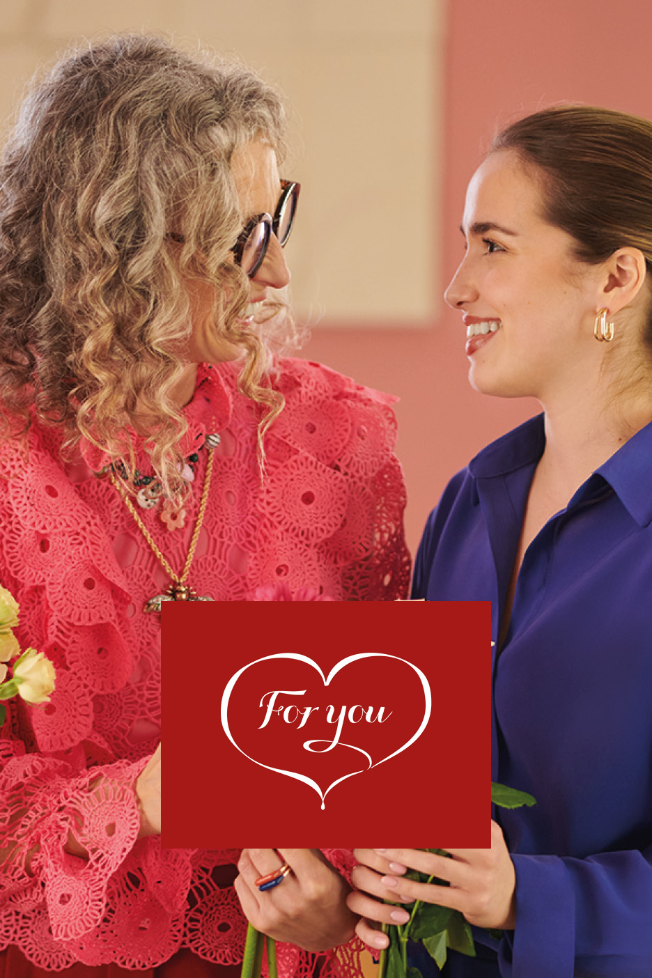 Due donne sorridenti si scambiano fiori e un biglietto rosso con la scritta 'For you'.